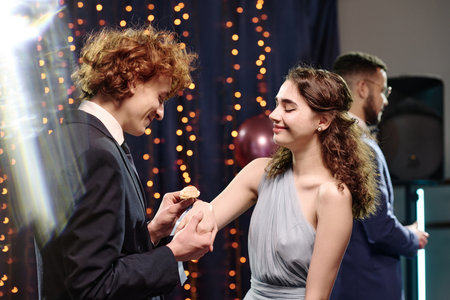 Caucasian teenage boy holding hand of Caucasian teenage girl while smiling at each other during formal event, another young adult man standing in background facing awayの写真素材