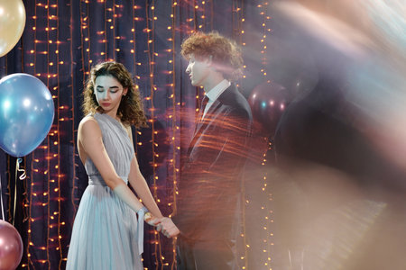 Teenage Caucasian girl holding hands with teenage Caucasian boy while standing together at dance party, both wearing formal attire, girl looking down, boy looking awayの写真素材