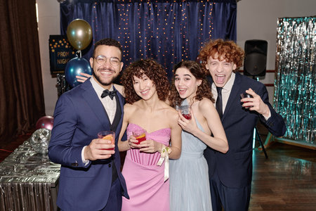 Group of multiethnic teenagers smiling and posing together while holding drinks at prom party, two...