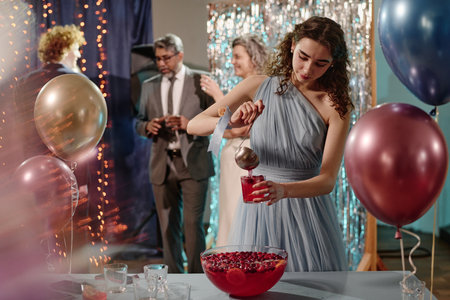 Teen in pastel blue dress serving punch from large bowl at festive party, standing near table with balloons while group of middle aged and senior adults socializing in backgroundの写真素材