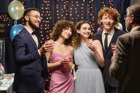 Group of multiethnic teenagers smiling and holding drinks while standing together at formal event,...