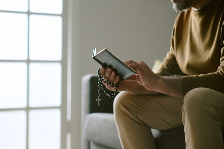 Middle aged Caucasian man sitting on sofa holding rosary beads and prayer book, reading quietly with focus on hands and religious objects, natural light coming from windowの写真素材
