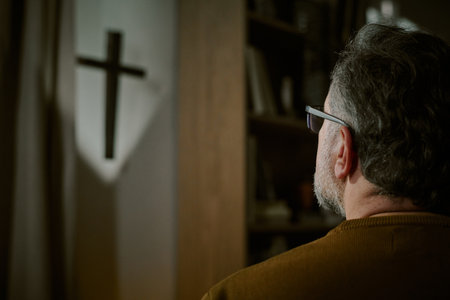 Middle aged Caucasian man with short hair and glasses standing indoors facing wall mounted cross, appearing contemplative, religious symbol casting shadow on wallの写真素材