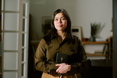 Portrait of young adult Asian woman standing indoors holding closed black book with cross on cover, looking directly at camera with neutral expression, bookshelf in backgroundの写真素材