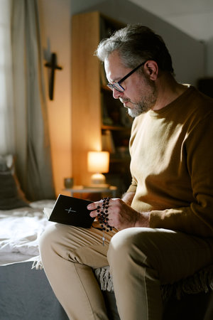 Middle aged Caucasian man sitting on bed reading religious book while holding rosary beads, wearing eyeglasses, focusing on spiritual practice in softly lit bedroomの写真素材