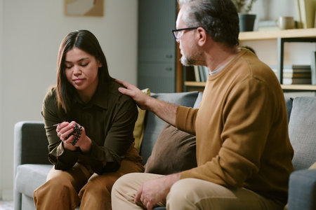 Middle aged Caucasian man comforting young adult Asian woman sitting on sofa, man placing hand on...