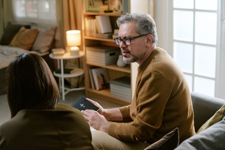 Middle aged Caucasian man wearing glasses holding Bible while talking to woman in living room, man sitting on sofa facing woman, bookshelf and lamp in backgroundの写真素材