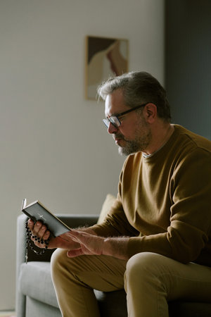 Middle aged Caucasian man sitting on sofa reading book and holding rosary beads, wearing glasses, appearing focused and contemplative, natural indoor setting with soft lightの写真素材