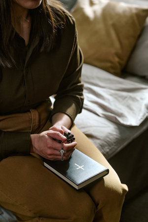 Young adult woman sitting on couch clasping hands in prayer holding rosary with cross, closed Bible...