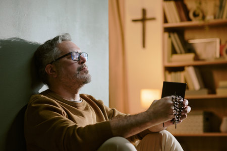 Middle aged Caucasian man sitting against wall holding rosary beads and prayer book, closing eyes in contemplation, bookshelf and cross visible in background, wearing glassesの写真素材