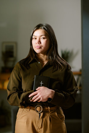 Portrait of young adult Asian woman standing indoors holding closed Bible with both hands, looking directly at camera with calm expression, long dark hair framing faceの写真素材