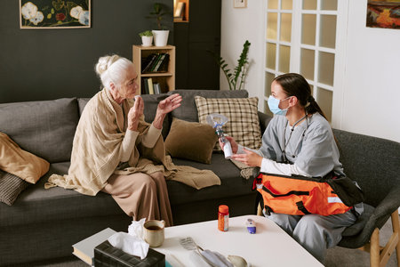 Senior Caucasian woman sitting on sofa gesturing while young adult Caucasian female nurse wearing medical mask kneeling and holding oxygen mask, medical supplies on table in foregroundの写真素材