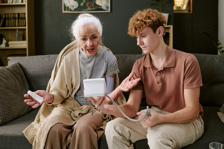 Senior Caucasian woman gesturing while young Caucasian male teenager measuring her blood pressure...