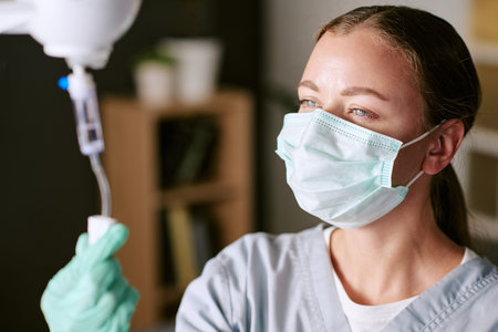 Caucasian young adult woman wearing medical mask and gloves adjusting intravenous drip in healthcare setting, focused expression visible, professional nurse performing medical procedureの写真素材