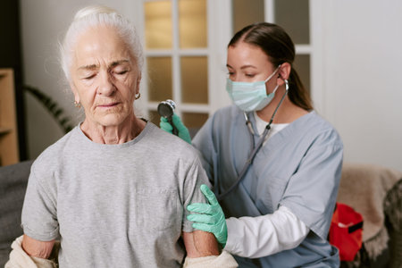 Senior Caucasian woman sitting with eyes closed while young adult Caucasian female nurse, wearing...