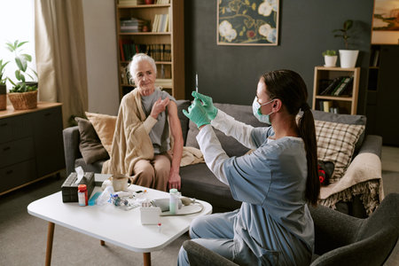 Senior Caucasian woman sitting on sofa preparing for vaccination while young adult Caucasian female nurse wearing mask and gloves, holding syringe in living room settingの写真素材