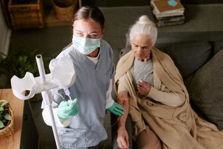 Caucasian female nurse wearing medical mask preparing intravenous drip for senior Caucasian woman sitting on sofa receiving treatment, nurse focusing on equipment while patient waitingの写真素材