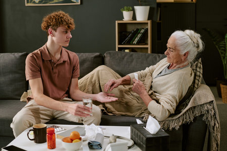 Teenage Caucasian boy giving glass of water and medication to senior Caucasian woman lying on sofa,...