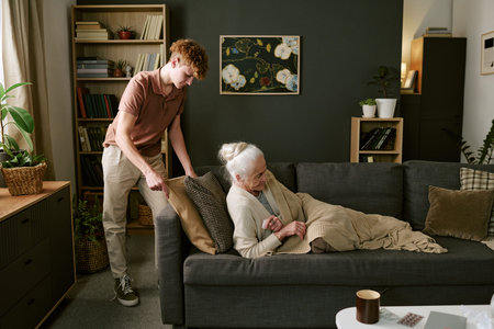 Caucasian teenage boy assisting senior Caucasian woman lying on sofa, holding pillow and blanket, interacting in living room setting with bookshelves and plants visible in backgroundの写真素材