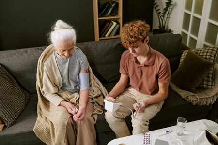 Senior Caucasian woman sitting on sofa having blood pressure measured by teenage Caucasian boy using digital monitor, both focused on health check, medication and water on tableの写真素材