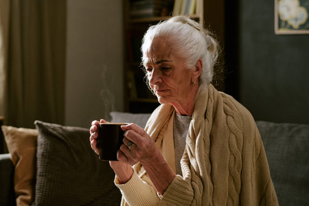 Senior Caucasian woman sitting on sofa holding mug with both hands, looking down thoughtfully,...