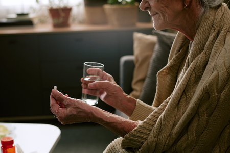 Senior Caucasian woman sitting indoors holding pill in one hand and glass of water in other hand,...