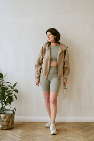 Vertical of young adult woman standing against plain wall holding water bottle, wearing activewear, looking to side, showing relaxed posture, short brown hair, indoor settingの写真素材