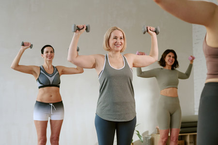 Group of middle aged Caucasian women exercising with dumbbells in fitness class, standing with arms raised and smiling, following instructor, engaging in strength training routineの写真素材