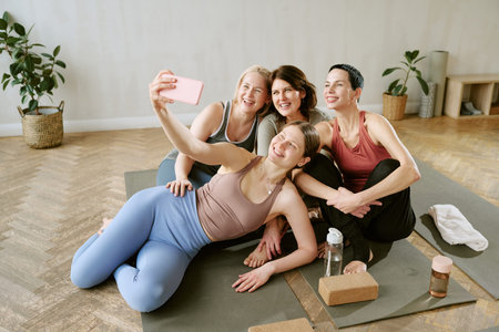 Group of four middle aged women sitting on yoga mats smiling and posing for selfie using smartphone during fitness class in studio, yoga blocks and water bottles nearbyの写真素材