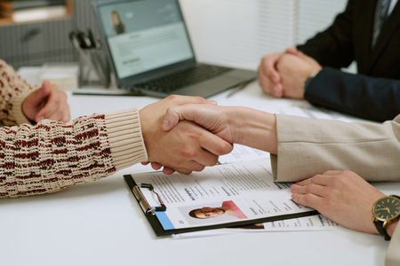 Woman and man shaking hands across desk during job interview, resume and clipboard visible on table,...