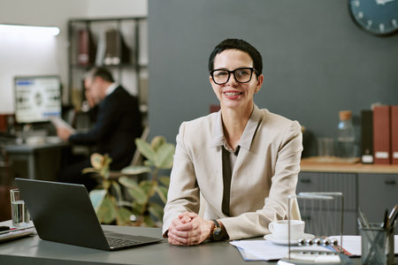 Portrait of middle aged Caucasian woman with short hair and glasses sitting at desk in modern office...