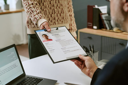 Caucasian middle aged man sitting at desk receiving clipboard with CV from young adult man standing...
