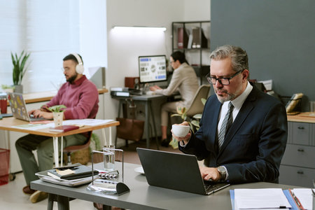 Middle aged Caucasian man wearing glasses working on laptop and drinking coffee in modern office,...