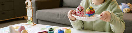 Website banner of child holding plate with colorful painted Easter eggs, sitting at table with art...