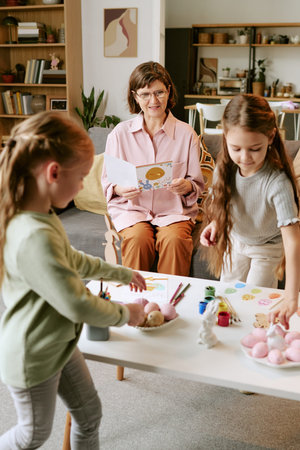 Middle aged woman sitting on sofa holding greeting card while two girls decorating eggs and crafting...