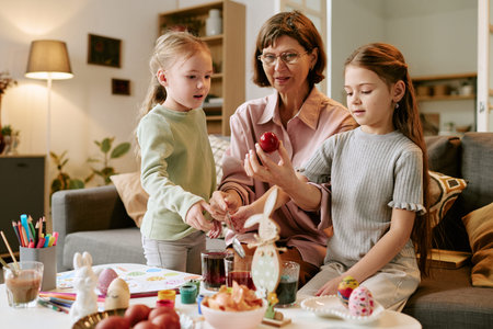 Middle aged Caucasian woman sitting with girls decorating Easter eggs together at table, children...