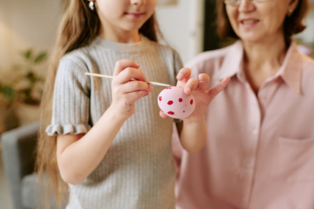 Child girl painting pink egg with brush while middle aged woman watching closely, both engaging in creative activity together indoorsの写真素材