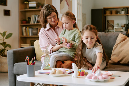 Middle aged woman sitting on sofa interacting with two girls, one girl holding pastel colored egg while other girl reaching for art supplies on tableの写真素材
