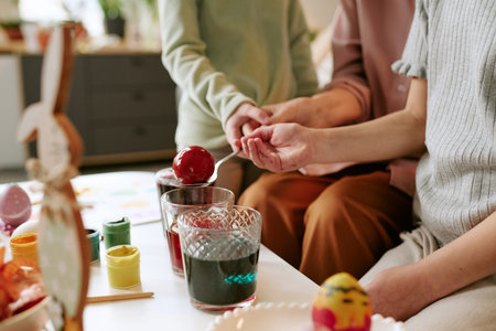 Caucasian child and woman dyeing Easter eggs together, holding spoon with red egg over glass of...