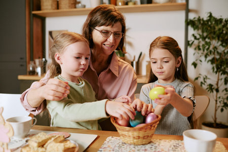 Caucasian woman sitting with two girls, smiling and helping children picking colorful eggs from...