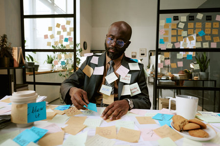 Black man sitting at cluttered desk covered with sticky notes, working with papers and reminders,...
