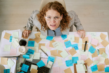 Portrait of Caucasian woman wearing eyeglasses sitting at cluttered office desk covered with sticky...