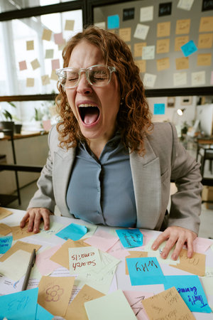 Caucasian young woman wearing glasses screaming with frustration while sitting at cluttered desk covered in sticky notes in modern office workspace, surrounded by business remindersの写真素材