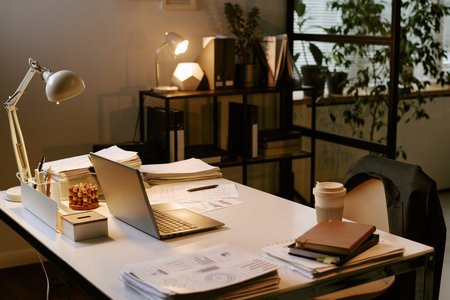 Modern office desk holding open laptop, documents, coffee cup, notebooks, pen holder, desk lamp and potted plants, business workspace arranged for professional work and productivityの写真素材
