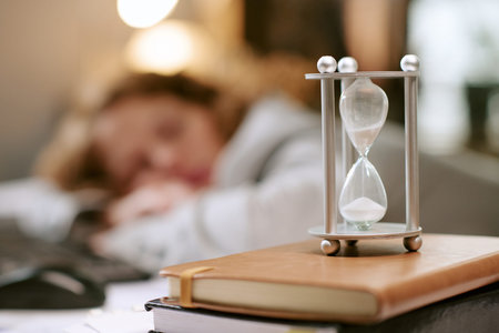 Caucasian young adult woman sleeping at desk in office with hourglass and notebook in foreground,...