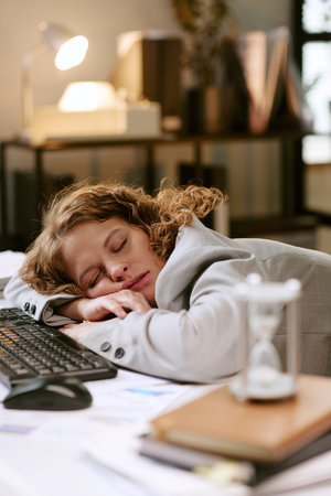 Young adult woman sleeping at desk, resting head on arms near computer keyboard surrounded by...