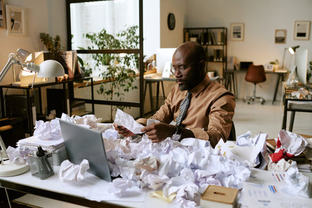 Black man sitting at cluttered desk surrounded by crumpled papers, working with documents and using laptop in modern office, appearing focused on business tasksの写真素材