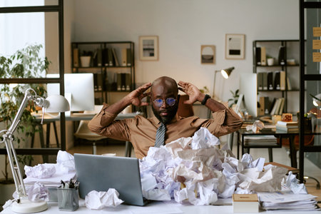 Black man sitting at cluttered desk surrounded by crumpled papers, holding head in hands, appearing stressed while working on laptop in modern business officeの写真素材