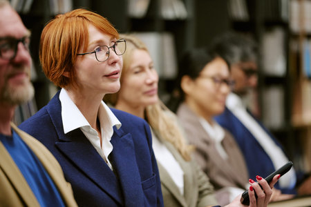 Caucasian middle aged woman with short red hair and glasses listening attentively during presentation, holding smartphone, sitting among diverse group of adults in book-filled settingの写真素材