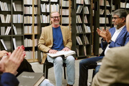 Middle aged Caucasian man sitting with open book on lap smiling during presentation in library, surrounded by diverse group of adults clapping and holding booksの写真素材